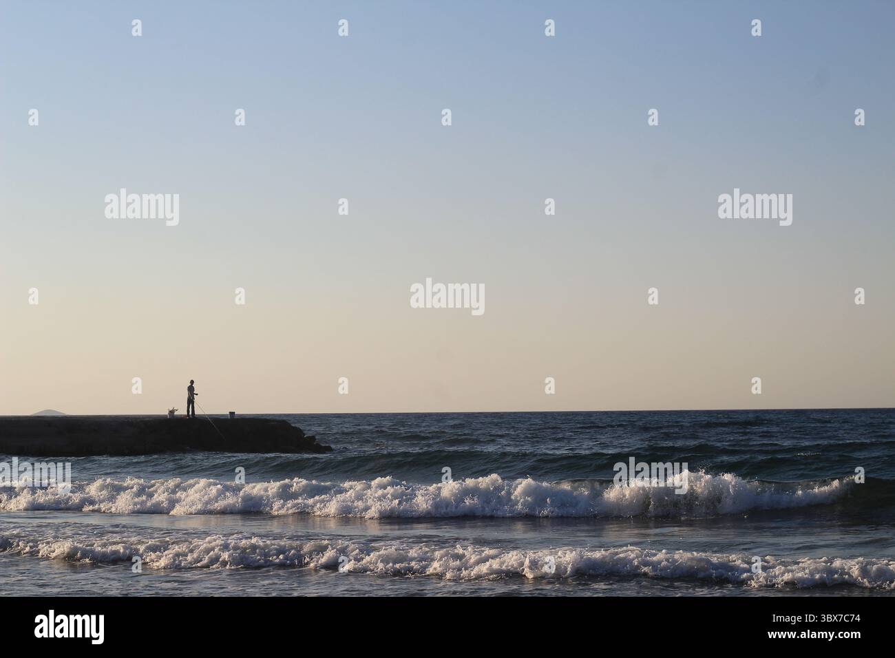 Une scène côtière tranquille au coucher du soleil met en scène un pêcheur solitaire debout sur une jetée rocheuse avec une canne à pêche. De douces vagues roulent sur le rivage sablonneux. Banque D'Images
