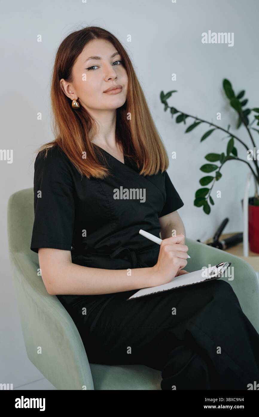 jeune femme esthéticienne vêtue de gommages médicaux noirs tenant une planche à pince tout en étant assise sur un fauteuil dans une clinique moderne Banque D'Images