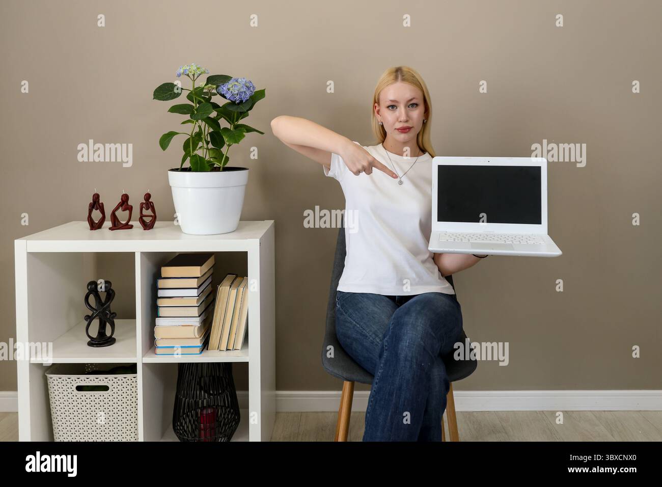 Jeune femme en tenue décontractée assise dans un intérieur élégant, tenant un ordinateur portable avec un écran vierge et pointant vers lui, suggérant la promotion ou le présentat Banque D'Images