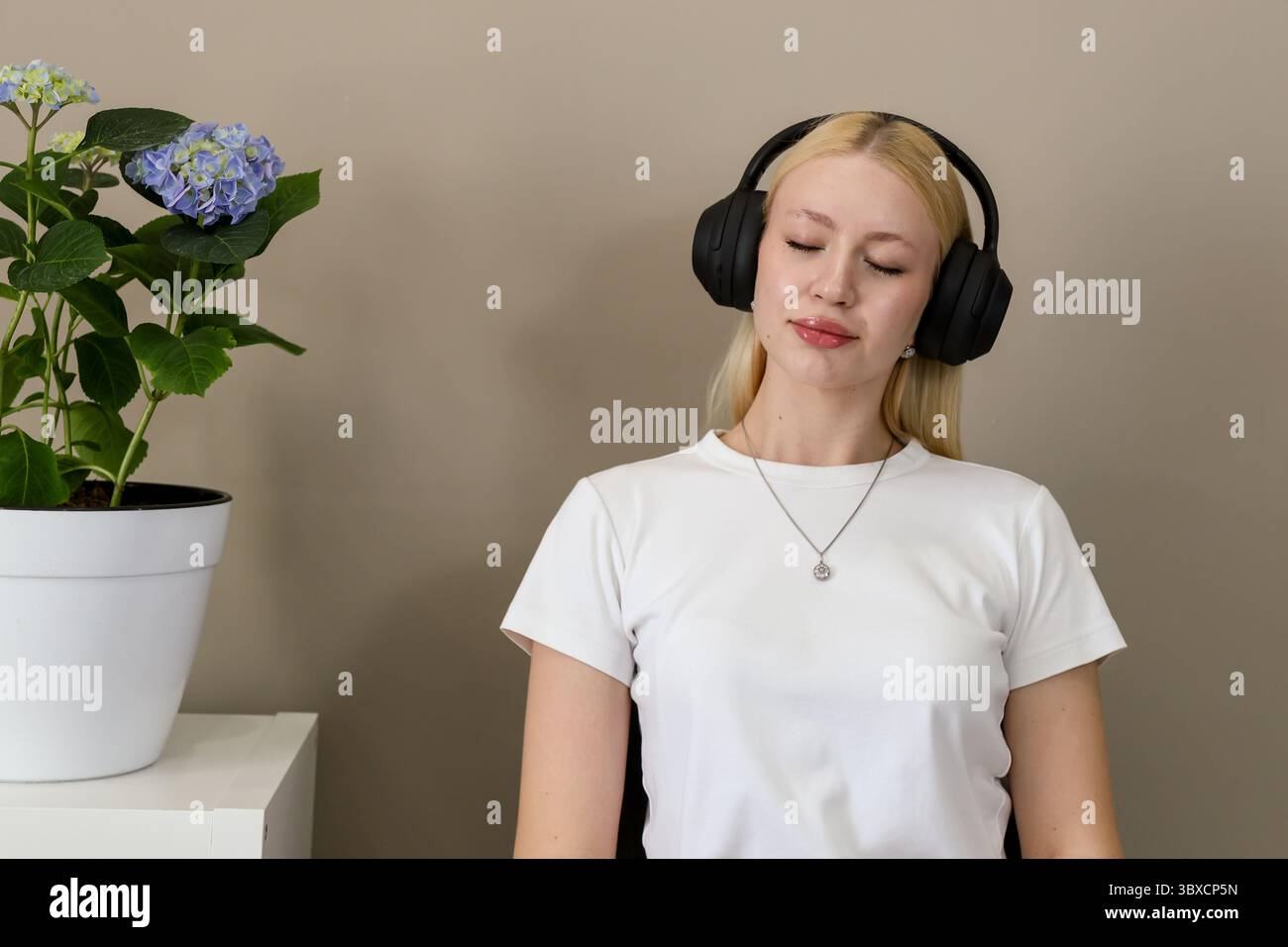 Jeune femme blonde dans un t-shirt blanc s'assoit paisiblement avec les yeux fermés, portant des écouteurs supra-auriculaires noirs et écoutant de la musique dans une pièce moderne et confortable Banque D'Images