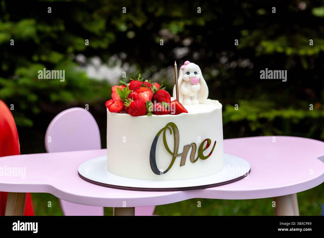 Gâteau blanc givré décoré de fraises fraîches et d'une jolie figurine de lapin, avec le mot One écrit sur le devant, placé sur une chaise haute rose ou Banque D'Images
