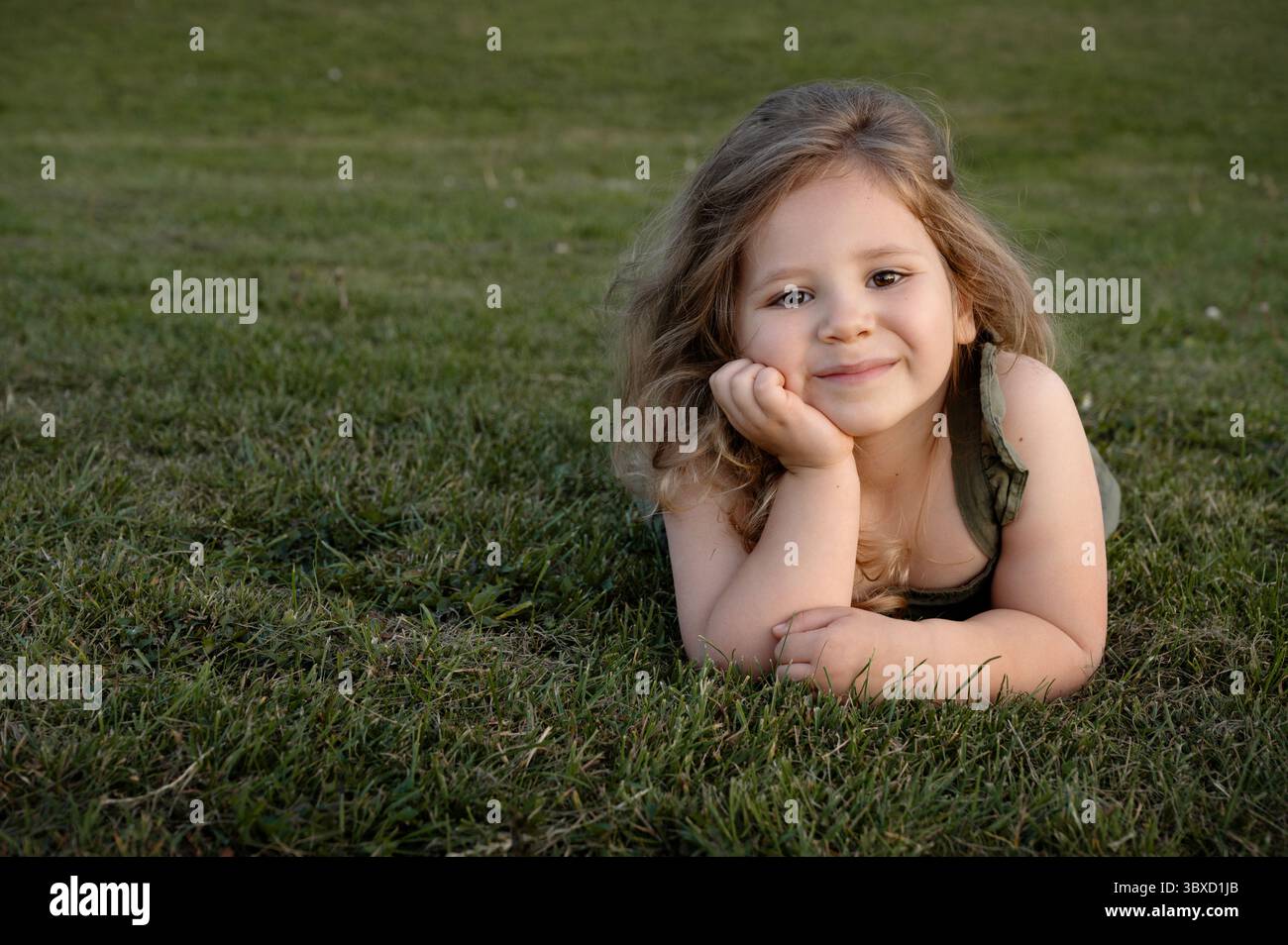 Fille enfant souriante couchée sur l'herbe, regardant la caméra. Menton sur les mains. Banque D'Images