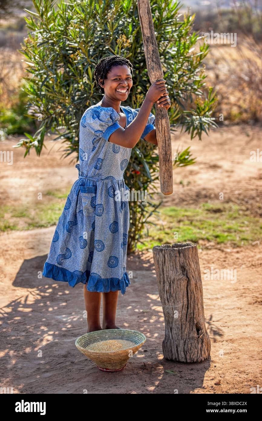 Village femme africaine emboutissant cuisine avec un pilon en bois dans un grand mortier pour préparer la farine broyant le sorgho de millet . Elle est habillée selon une tradition Banque D'Images