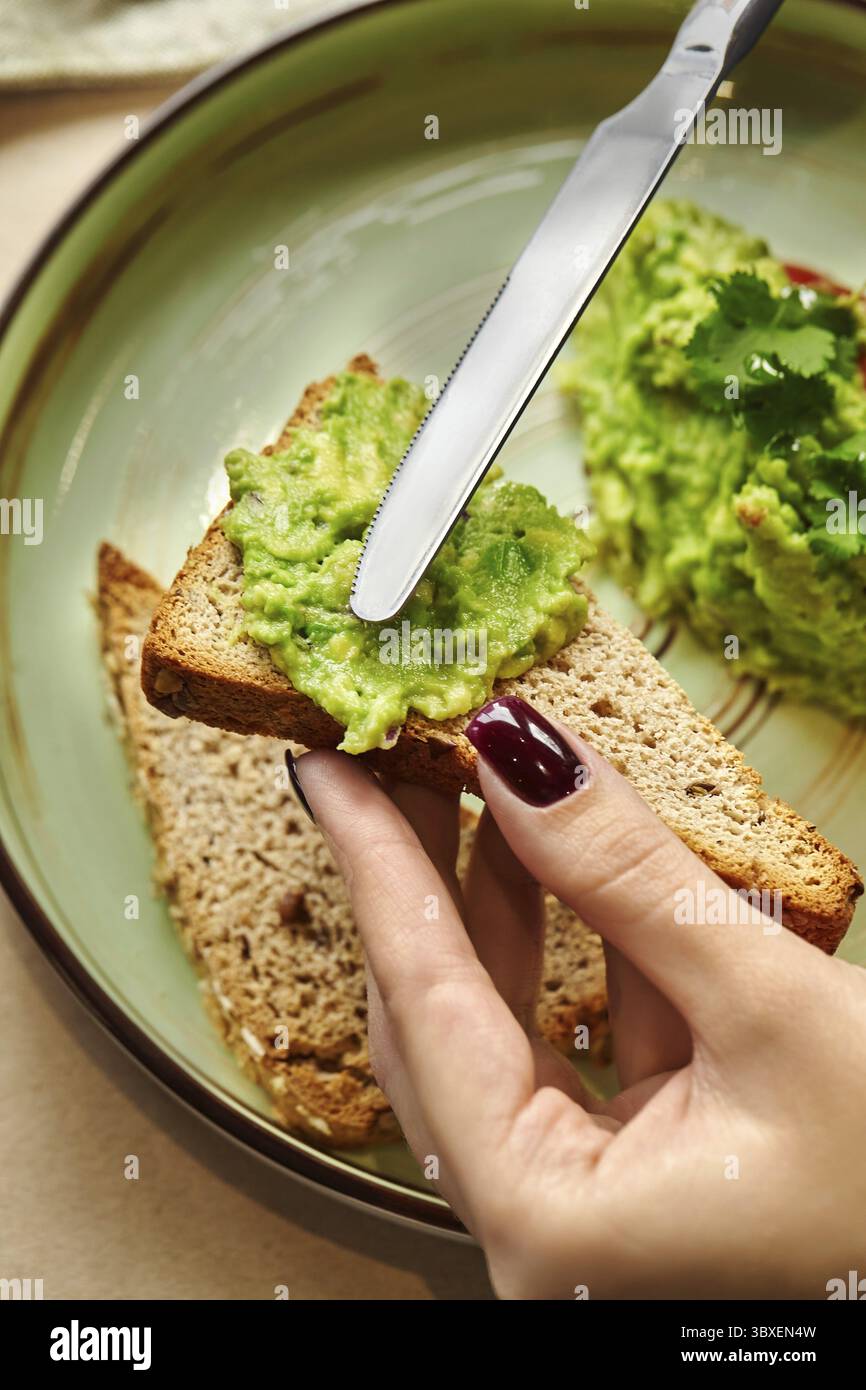 Main de femme étalant de la purée d'avocat sur une tranche de pain grillé de blé entier, préparant un petit déjeuner ou une collation sain et nutritif Banque D'Images