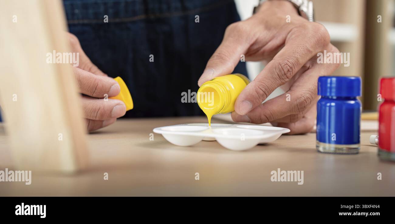 Mains versant de la peinture jaune dans une palette avec des bouteilles de peinture bleue et rouge sur une table en bois, prête pour un projet d'art. Banque D'Images