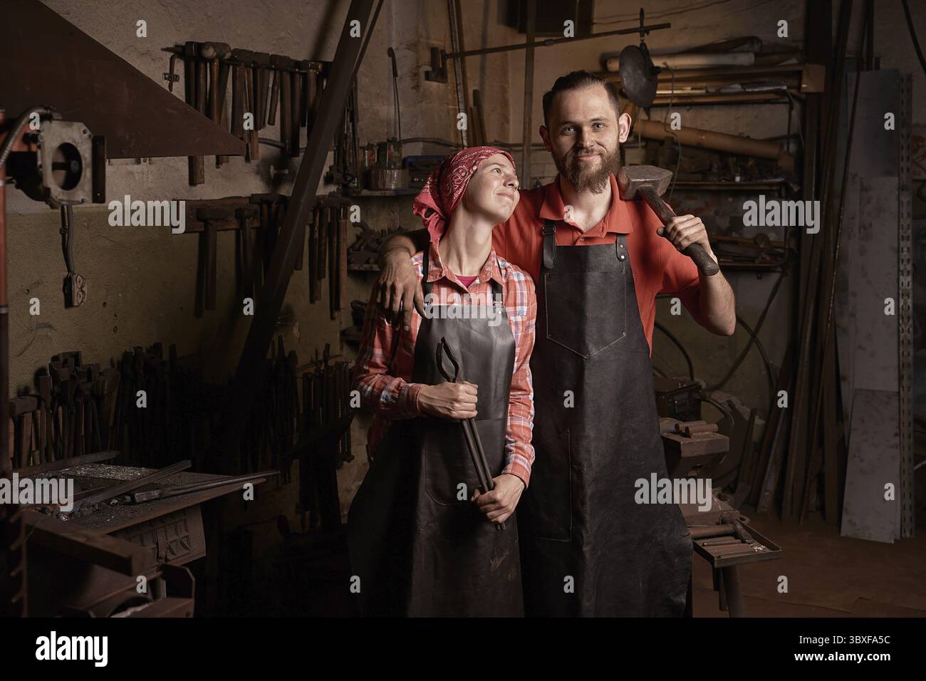 Forgerons professionnels en portrait de forgeron. Couple de jeune femme européenne souriante et homme barbu tiennent un marteau et une pince en métal. Travail en métal fait à la main i Banque D'Images