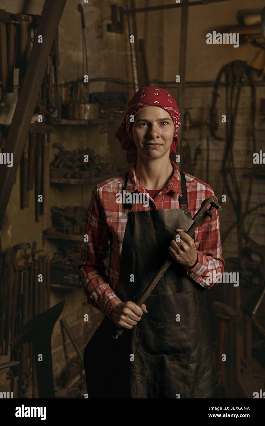 Forgeron professionnel en portrait d'atelier.Une jeune femme européenne souriante porte un uniforme et porte des détails en métal faits main.Concept d'artisanat Banque D'Images