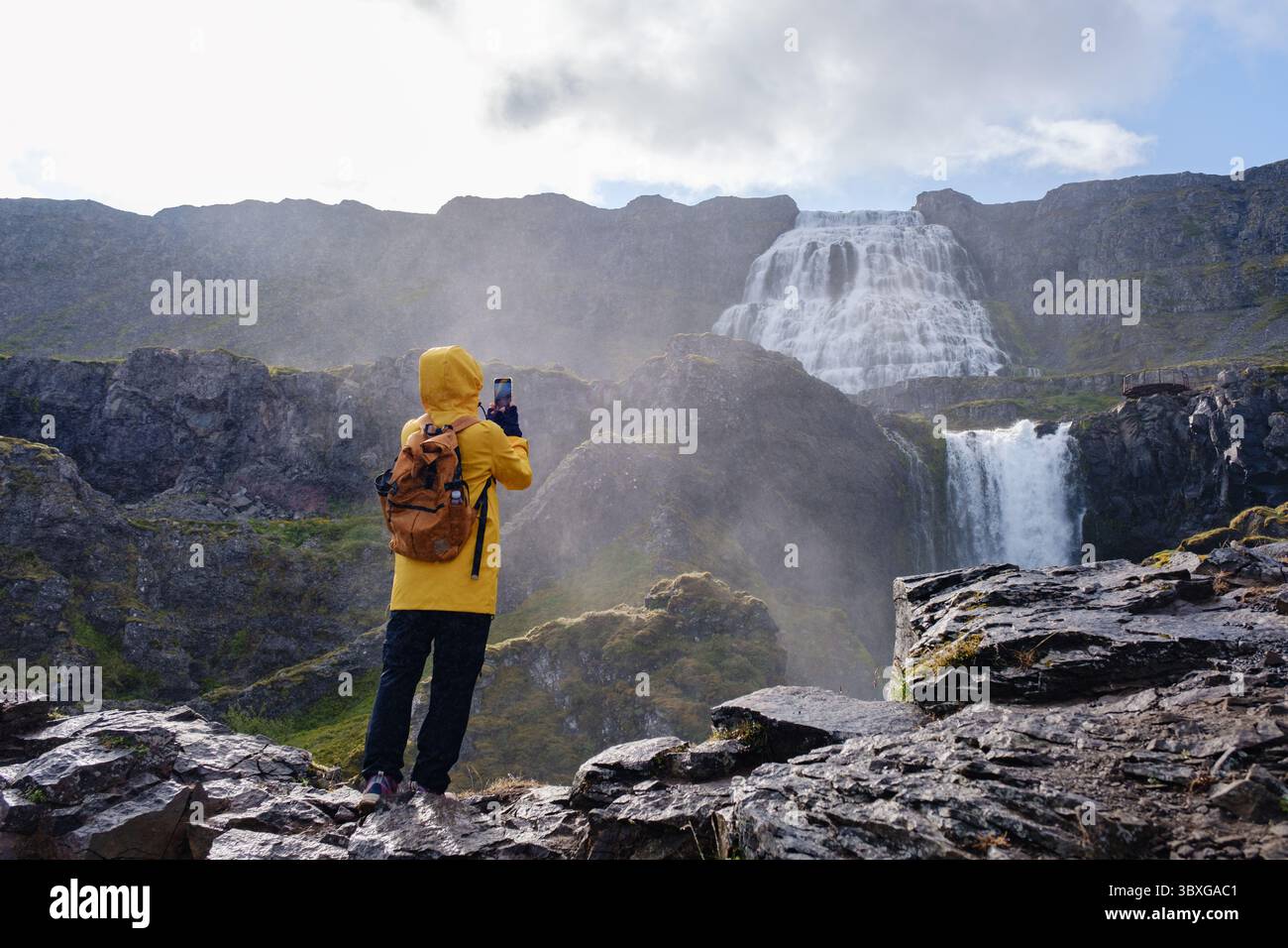 Un voyageur vêtu d'une veste de pluie jaune se tient sur un terrain rocheux, capturant la beauté de la cascade de Dynjandi, Westfjord, Islande. La cascade se déverse dramatiquement dans un paysage luxuriant sous un ciel nuageux Banque D'Images