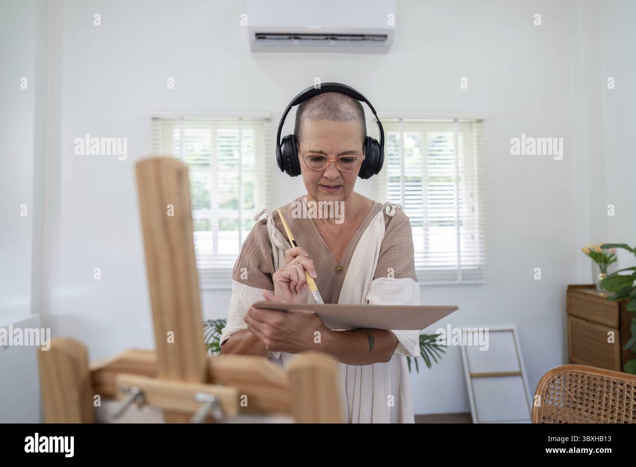 Femme âgée utilisant une tablette pour l'art numérique tout en portant des écouteurs dans un studio à domicile lumineux et moderne avec lumière naturelle Banque D'Images
