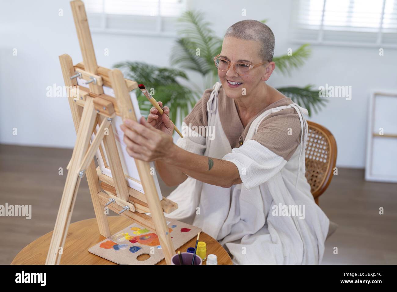 Artiste femme âgée peignant sur un chevalet dans une pièce bien éclairée, mettant en valeur la créativité et la passion pour l'art dans un environnement serein à la maison Banque D'Images