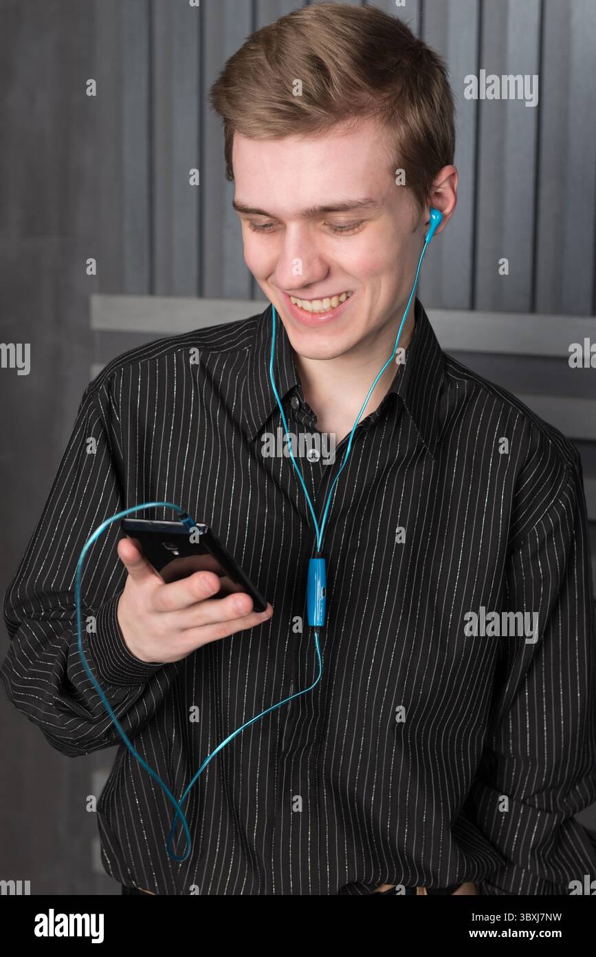 Un jeune homme dans une chemise noire écoutant de la musique de votre smartphone avec des écouteurs sur un fond sombre Banque D'Images