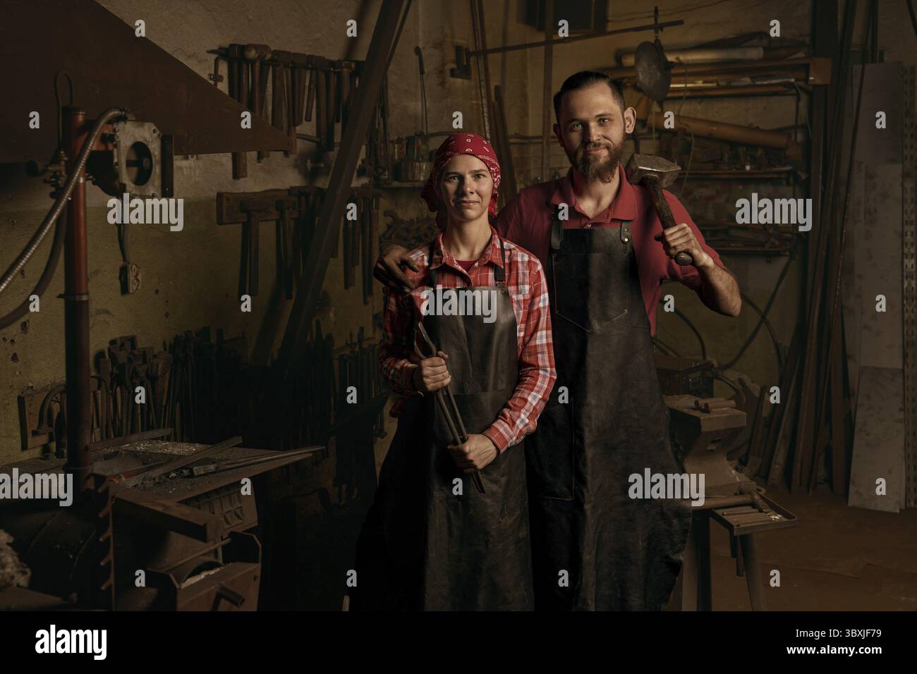 Forgerons professionnels en portrait de forgeron. Couple de jeune femme européenne souriante et homme barbu tiennent un marteau et une pince en métal. Travail en métal fait à la main i Banque D'Images