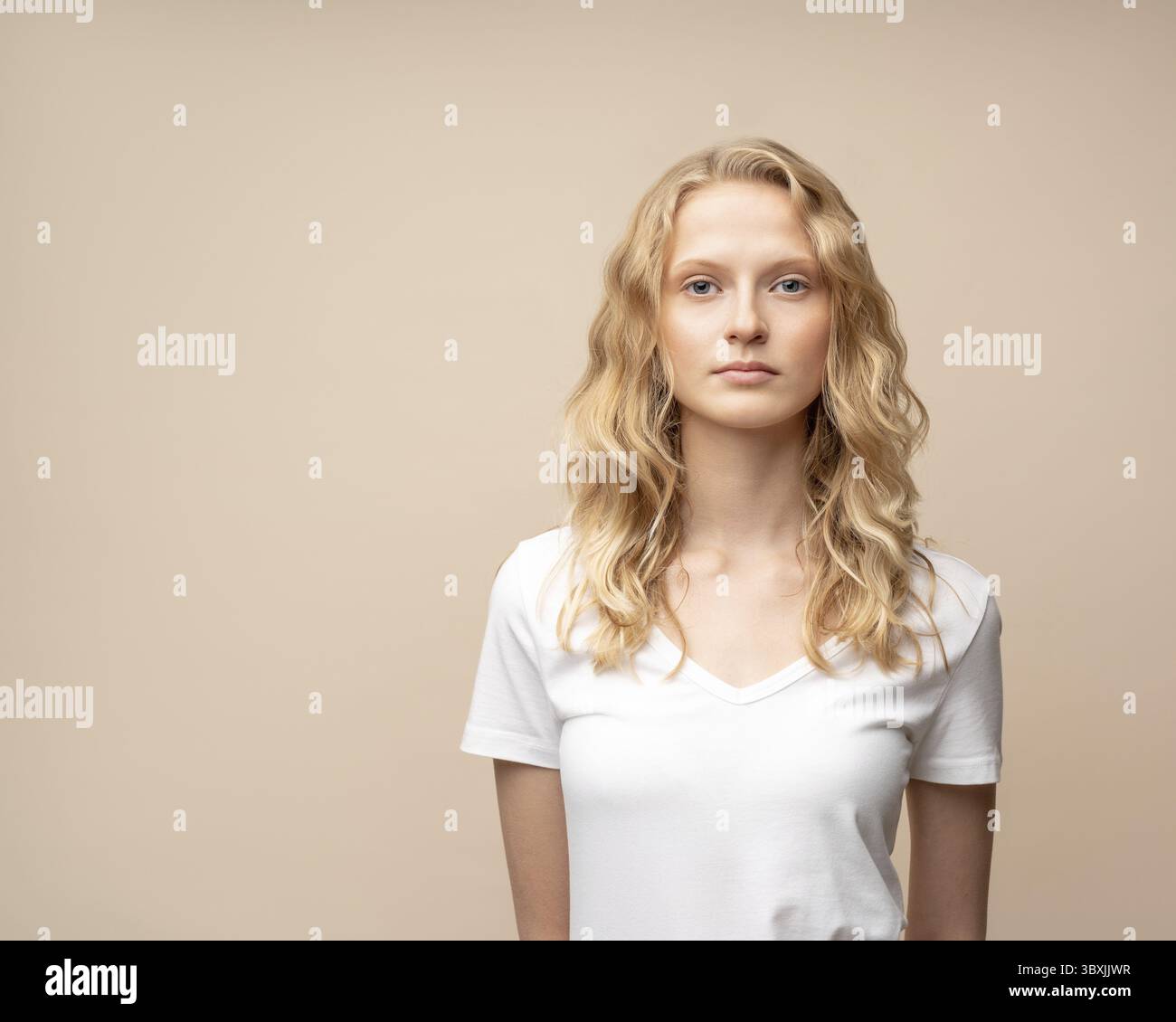 Portrait de taille haute de belle jeune femme blonde sérieuse intelligente sans maquillage sur le mur beige. Jolie femme avec des cheveux bouclés en t-shirt blanc Banque D'Images