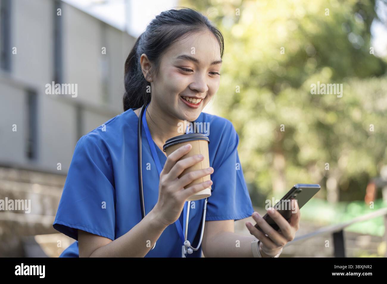 Une travailleuse de santé asiatique en gommage bleu sourit tout en dégustant un café et en utilisant son smartphone pendant une pause à l'extérieur Banque D'Images