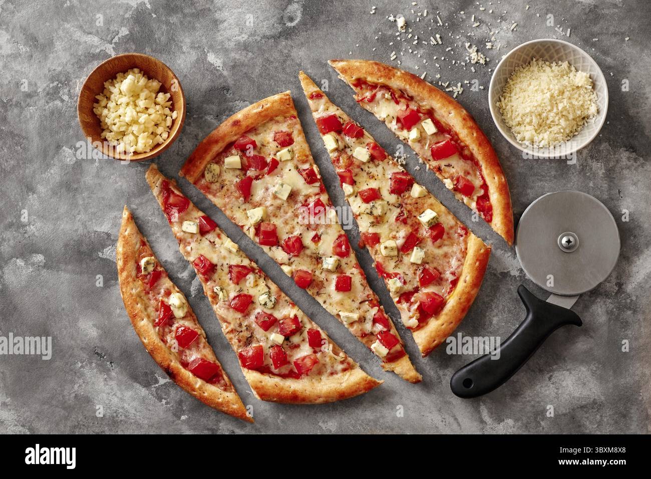 Pizza fine classique appétissante en tranches avec tomates fraîches juteuses, morceaux de fromage bleu et basilic séché sur une surface de pierre grise avec un couteau à roue et Banque D'Images