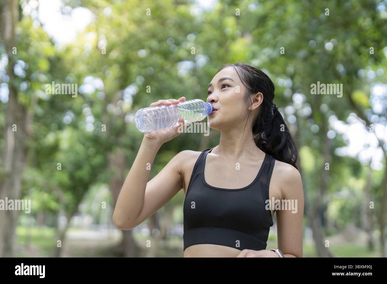 Une jeune femme en tenue athlétique boit de l'eau tout en prenant une pause de son entraînement dans un parc verdoyant luxuriant par une journée ensoleillée Banque D'Images