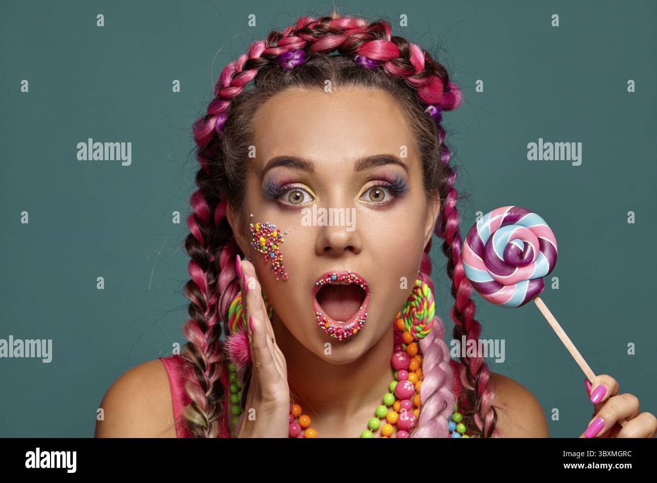 Photo en gros plan d'une magnifique fille avec une coiffure tresses multicolores et lumineux maquillage, posant en studio sur un fond bleu, tenant une loll Banque D'Images