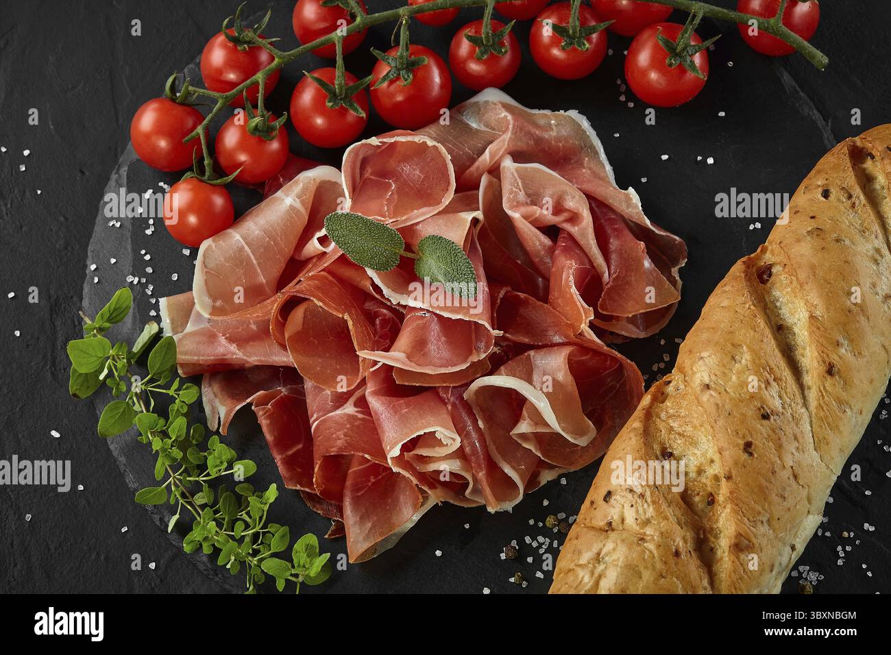 Délicieux, tranchés manuellement jamon, tomates cerises, baguette, fines herbes et épices sur ardoise noir bord contre un fond gris foncé. La cuisine, reste Banque D'Images