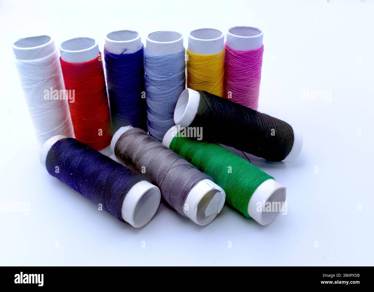 Dix bobines de fil à coudre vibrant en blanc, rouge, bleu, bleu clair, jaune, rose, noir, marron, violet et vert sont disposés sur une surface blanche. Banque D'Images