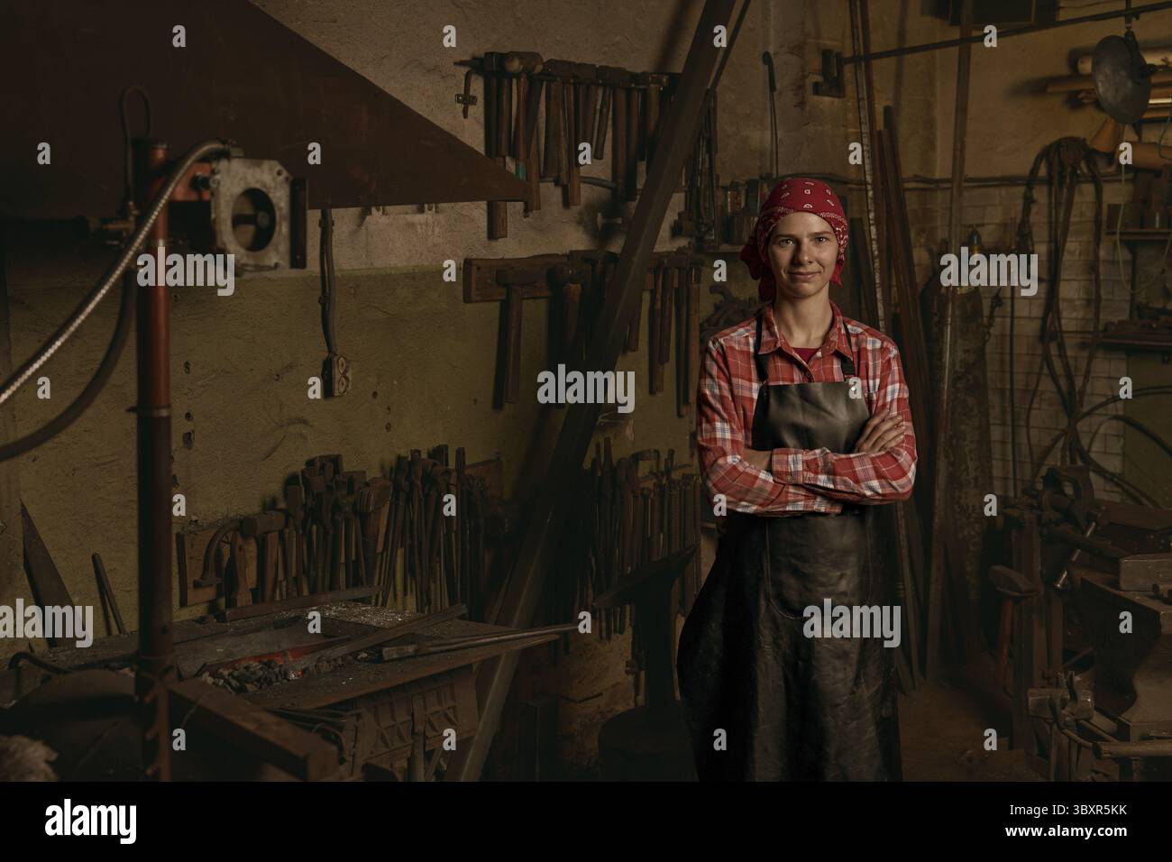 Forgeron professionnel en portrait forgeron. Jeune femme européenne souriante porte l'uniforme et les bras croisés. Idée de travail en métal fait à la main. Concept de handicap Banque D'Images