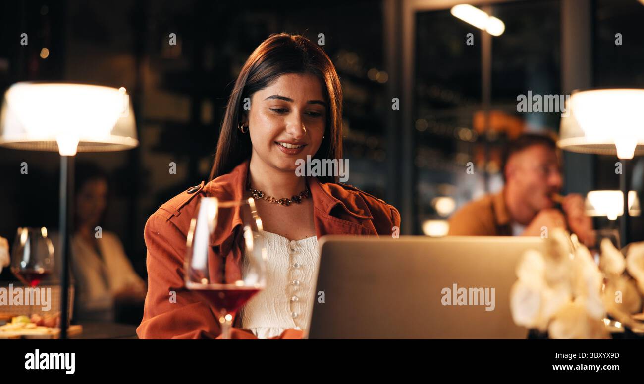 Ordinateur portable, vin et femme dans le restaurant la nuit avec blog en ligne pour l'examen de l'hospitalité avec freelance. Travail à distance, boisson et auteur féminin avec Banque D'Images