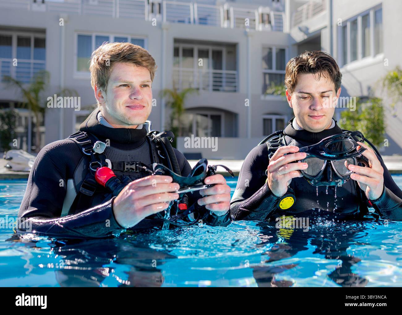 Deux plongeurs dans une piscine d'un centre de plongée rincent leurs masques de plongée dans l'eau, portant des combinaisons et de l'équipement complets. Banque D'Images