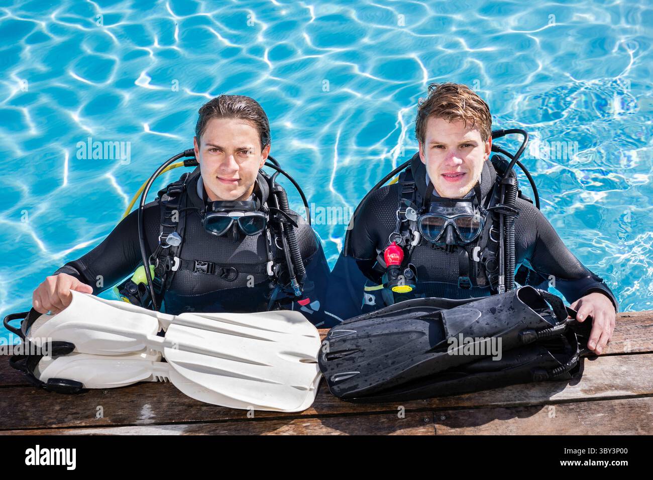 Deux plongeurs debout dans une piscine, regardant la caméra, souriant avec leurs palmes devant eux sur le pont. Port de combinaisons complètes et d'équipement de plongée Banque D'Images
