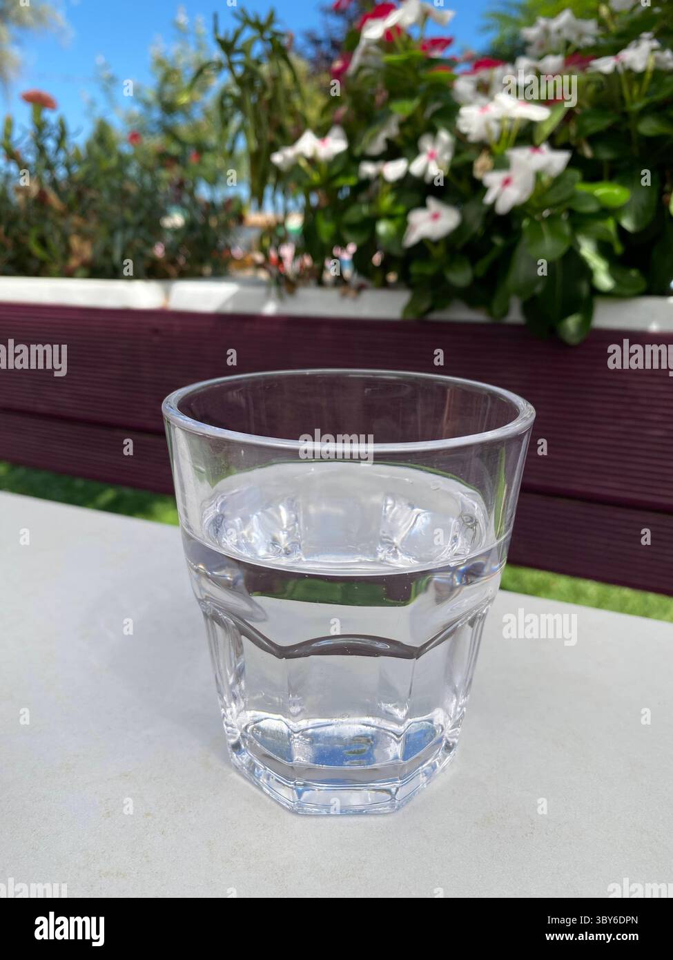 Verre transparent à facettes rempli d'eau, placé sur une table extérieure de couleur claire. L'eau à l'intérieur apparaît immobile et claire, reflétant un peu de lumière. Banque D'Images