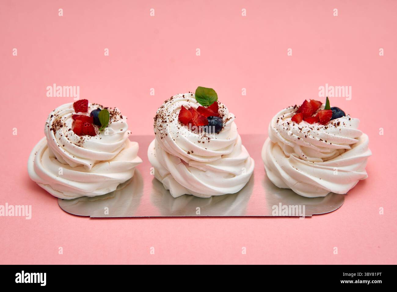 Trois meringues élégamment plaquées surmontées de baies mélangées et de menthe créent un dessert visuellement délicieux sur un fond rose doux, mettant en valeur leur Banque D'Images