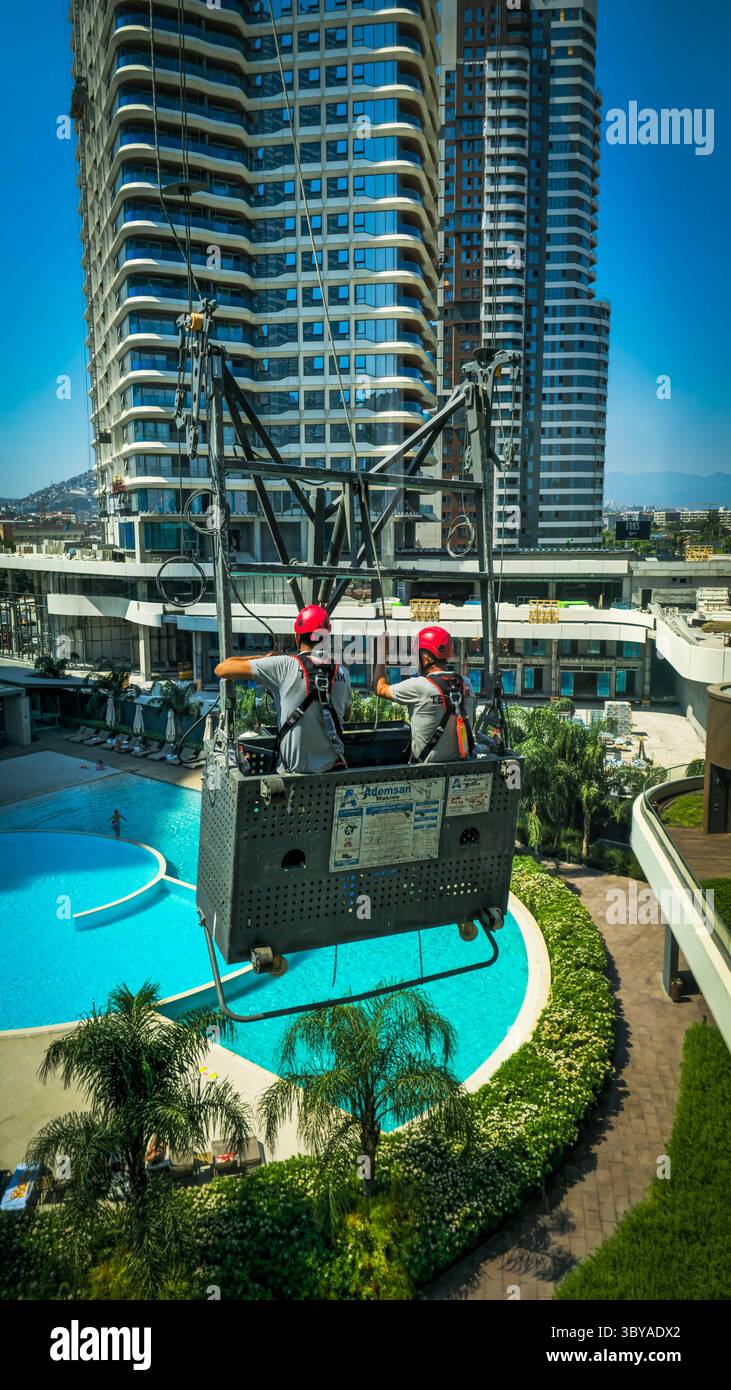 Deux ouvriers de la construction travaillent à l'extérieur deux ouvriers de la construction portant des casques et des harnais de sécurité se tiennent sur un berceau suspendu dans un immeuble de grande hauteur, travaillant à l'extérieur par une journée ensoleillée avec un paysage urbain en arrière-plan Izmir Konak Turquie Copyright : xx 20250719_111922 Banque D'Images