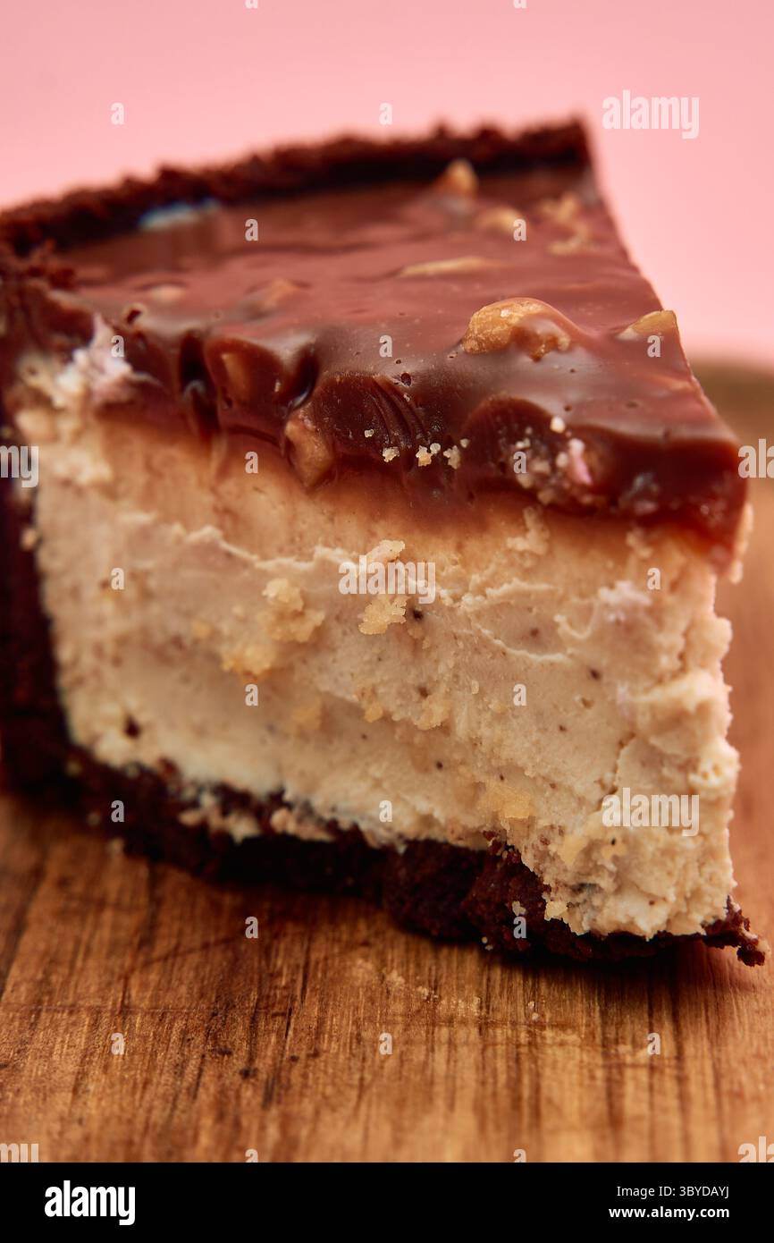 Une délicieuse tranche de cheesecake au beurre de cacahuète au chocolat garnie d'une riche ganache, présentée sur une planche de bois sur un fond rose. Banque D'Images