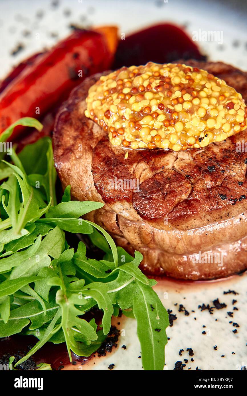 Un succulent steak de bœuf grillé garni d'une généreuse garniture de moutarde est servi avec des feuilles de roquette fraîches et des poivrons rouges carbonisés, créant ainsi un vi Banque D'Images