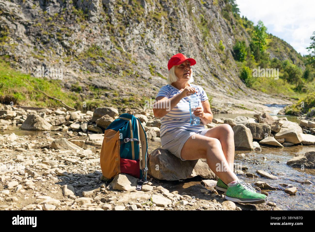 Une femme âgée portant une casquette rouge et une chemise rayée est assise sur un rocher dans un ruisseau de montagne rocheux, profitant d'un verre d'eau pendant une pause randonnée. Son backpa Banque D'Images