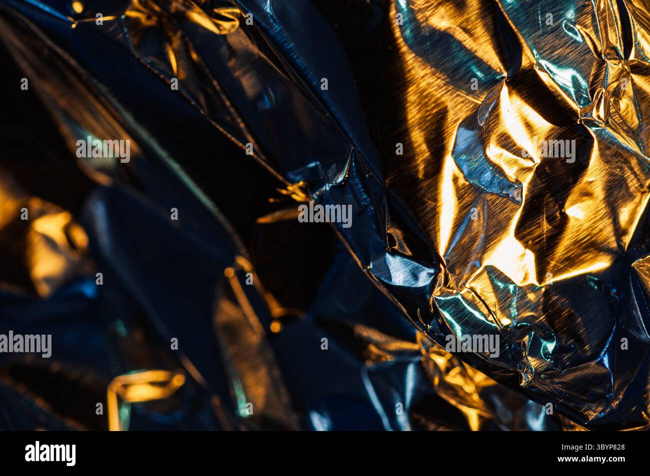Gros plan abstrait d'une feuille métallique froissée, reflétant un mélange de lumière chaude dorée et bleue froide. Banque D'Images