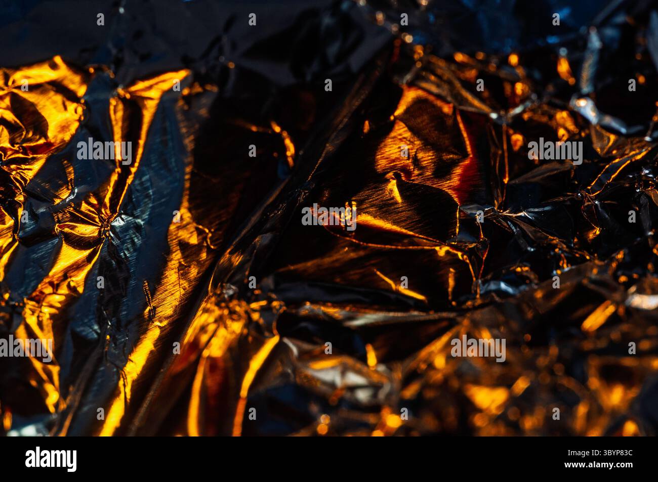 Gros plan d'une feuille d'aluminium froissée reflétant une lumière chaude et dorée. Les ombres et les reflets contrastés créent une composition dynamique et abstraite. Banque D'Images