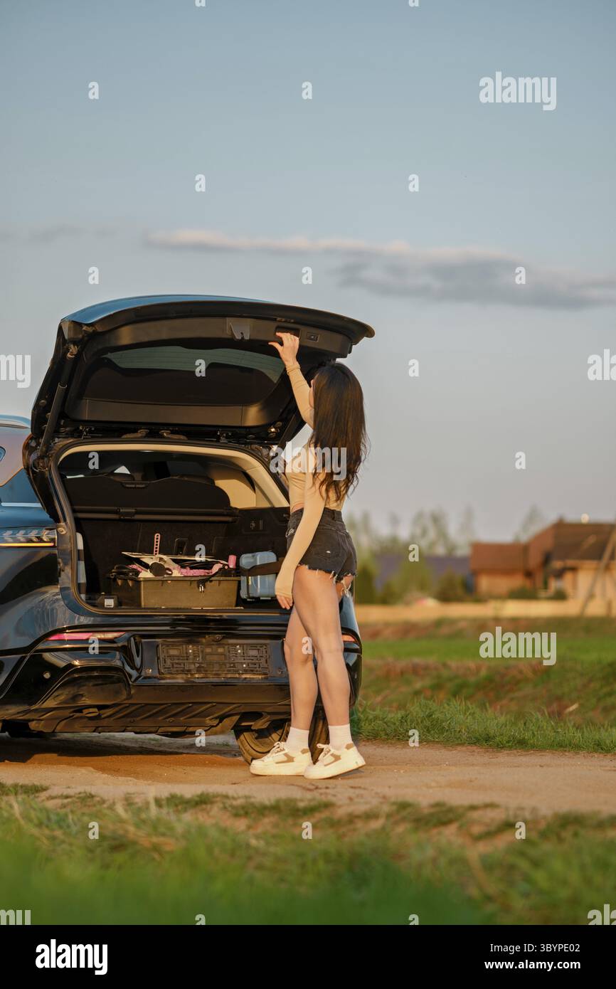 Une femme se tient à côté d'une voiture garée dans un champ ouvert, soulevant le coffre pour accéder à l'équipement d'aventure. Le ciel lumineux et le paysage vert créent un s parfait Banque D'Images Une femme se tient à côté d'une voiture garée dans un champ ouvert, soulevant le coffre pour accéder à l'équipement d'aventure. Le ciel lumineux et le paysage vert créent un s parfait Banque D'Images