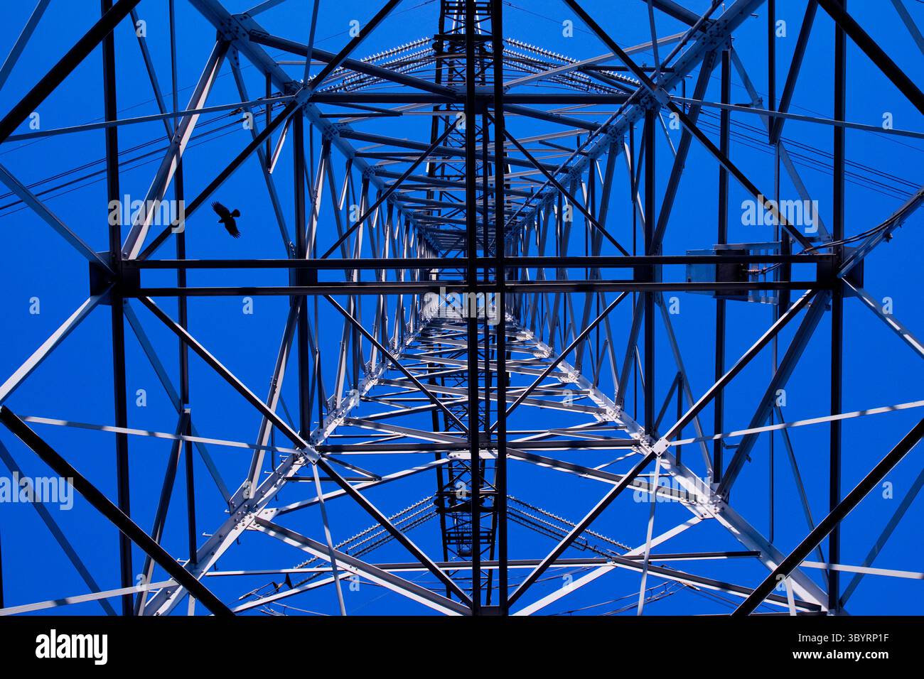 Perspective spectaculaire à faible angle d'une tour de transmission haute tension en treillis d'acier contre un ciel bleu vif, mettant en valeur la géométrie industrielle et la puissance. Banque D'Images