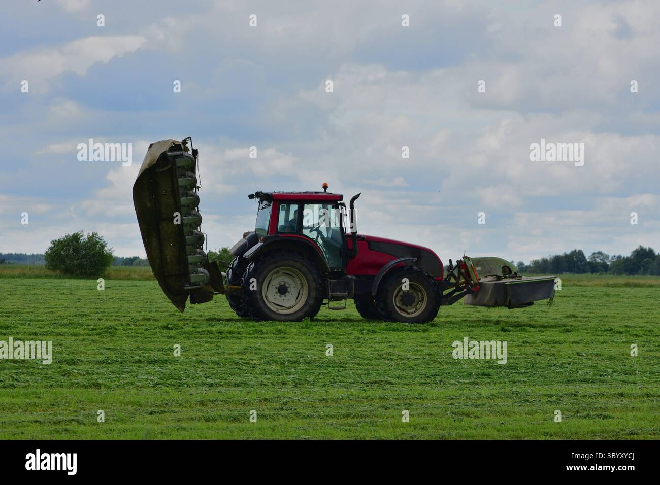 Vue latérale d'un tracteur rouge avec équipement de fauche relevé sur un terrain herbeux sous un ciel nuageux. Machines agricoles pendant le travail saisonnier. Banque D'Images