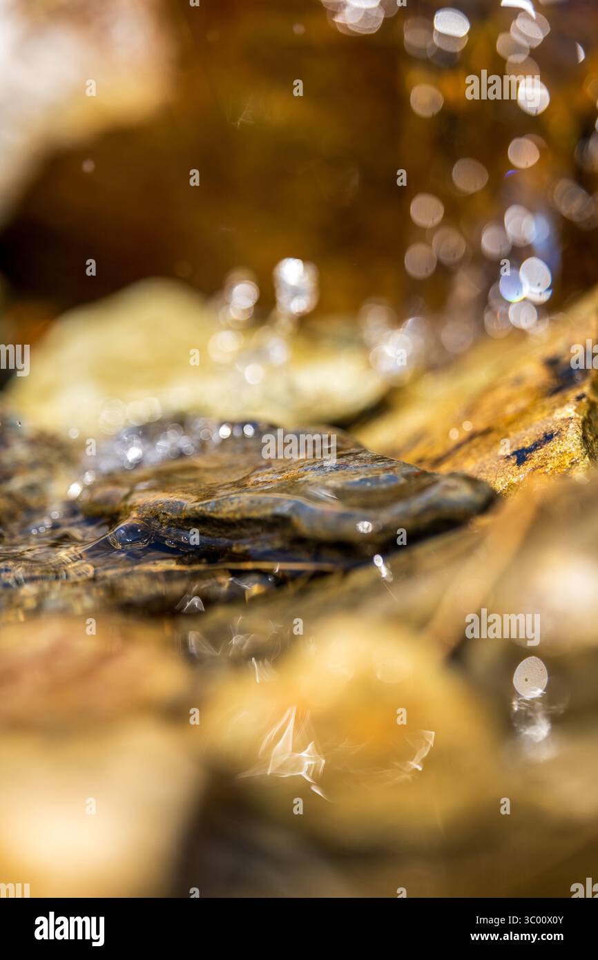 Des gouttelettes d'eau étincelantes coulent sur des pierres de ruisseau en couches, leurs surfaces altérées et leur bokeh ambré révélant un mouvement fluide dans le microcosme Banque D'Images