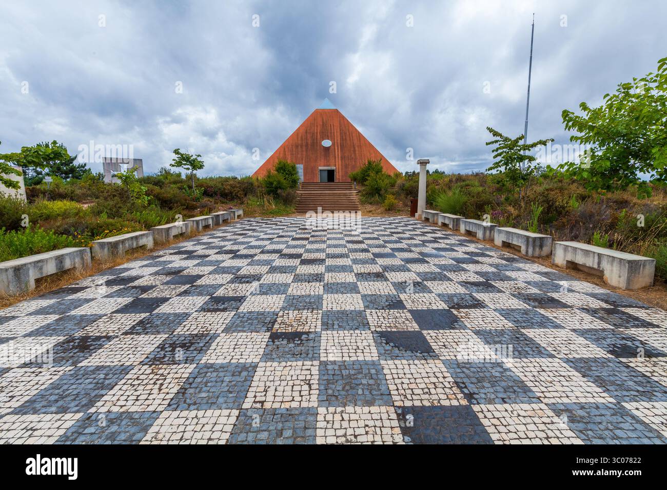 Une impressionnante pyramide rouge se dresse au bout d'un sentier en mosaïque à carreaux, encadré de verdure et menant à un cadre extérieur serein sous un ciel nuageux Banque D'Images