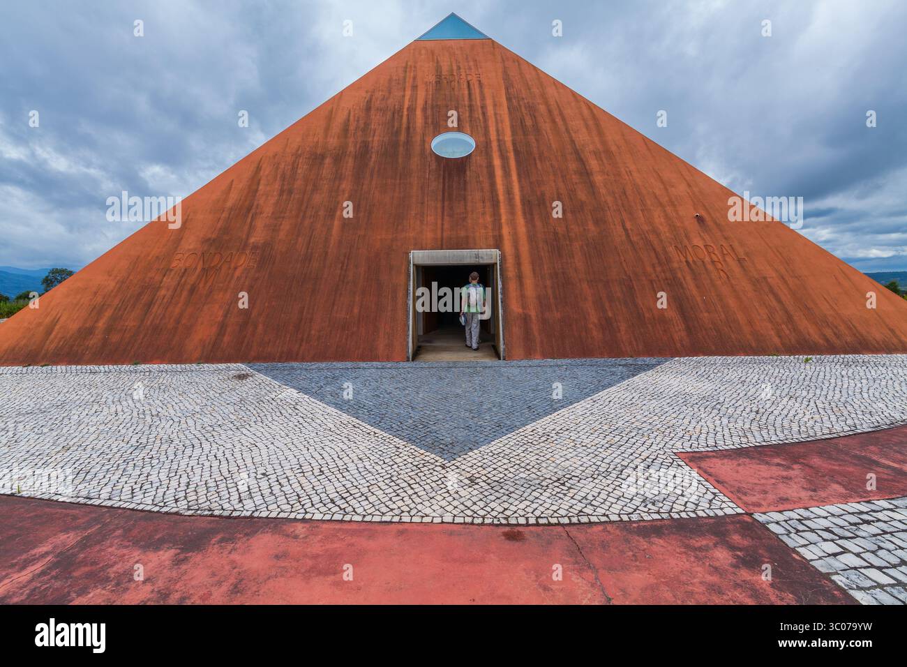 Une structure en forme de pyramide rouge frappante avec des inscriptions se tient sous un ciel nuageux, alors qu'une personne entre dans son entrée par un jour nuageux Banque D'Images