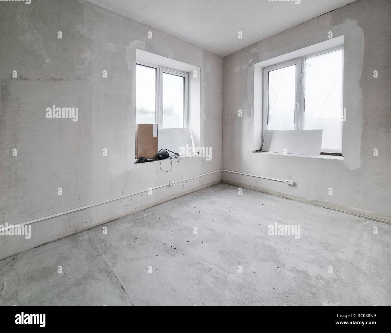 Rénovation intérieure de la maison ou construction inachevée. Pièce vide avec deux grandes fenêtres et murs blancs non peints. Banque D'Images