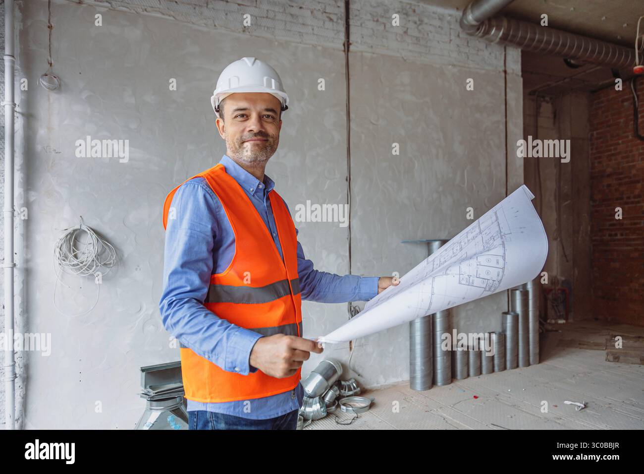 Un directeur de la construction portant un gilet orange et un casque blanc tient des plans tout en se tenant debout dans un chantier de rénovation avec des murs de briques et du métal exposés Banque D'Images
