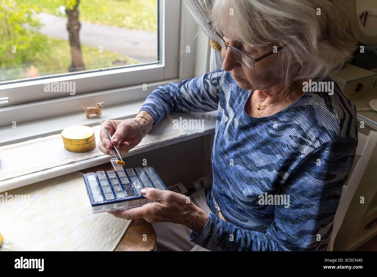 Une femme âgée aux cheveux gris organise méticuleusement ses pilules à l'aide d'une pince à épiler, assise près de la fenêtre dans le confort de sa propre maison. Banque D'Images