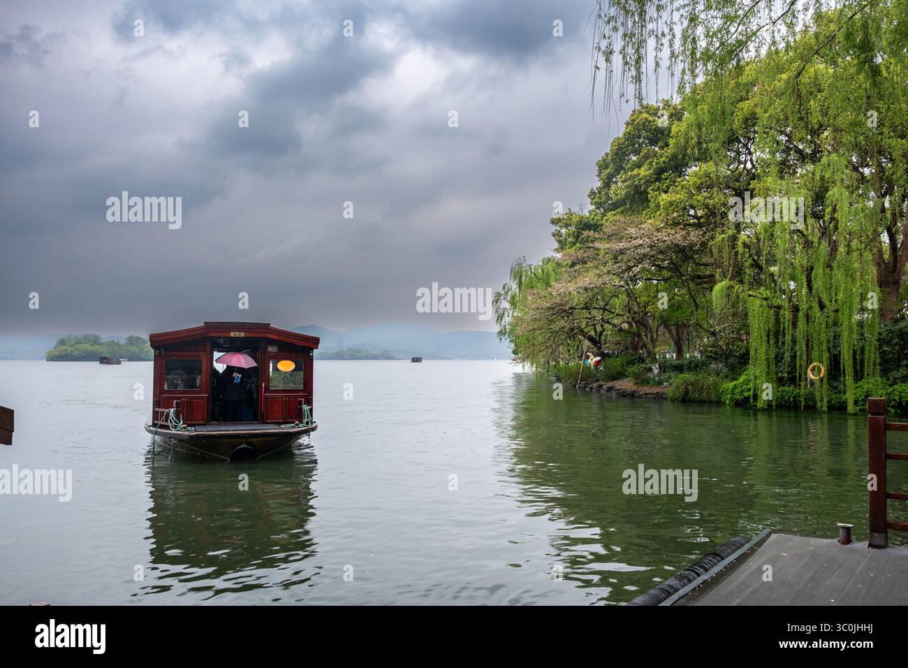 Après-midi pluvieux au lac de l'Ouest, Hangzhou, Chine. Bateau traditionnel en bois approche du quai. Saules verdoyants et collines brumeuses Banque D'Images