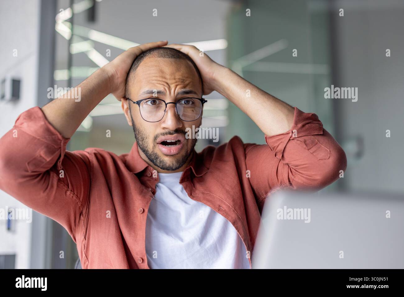 Un jeune homme inquiet avec des lunettes réagit à ce qu'il voit sur son ordinateur portable, se tenant la tête dans un état inquiet. Banque D'Images