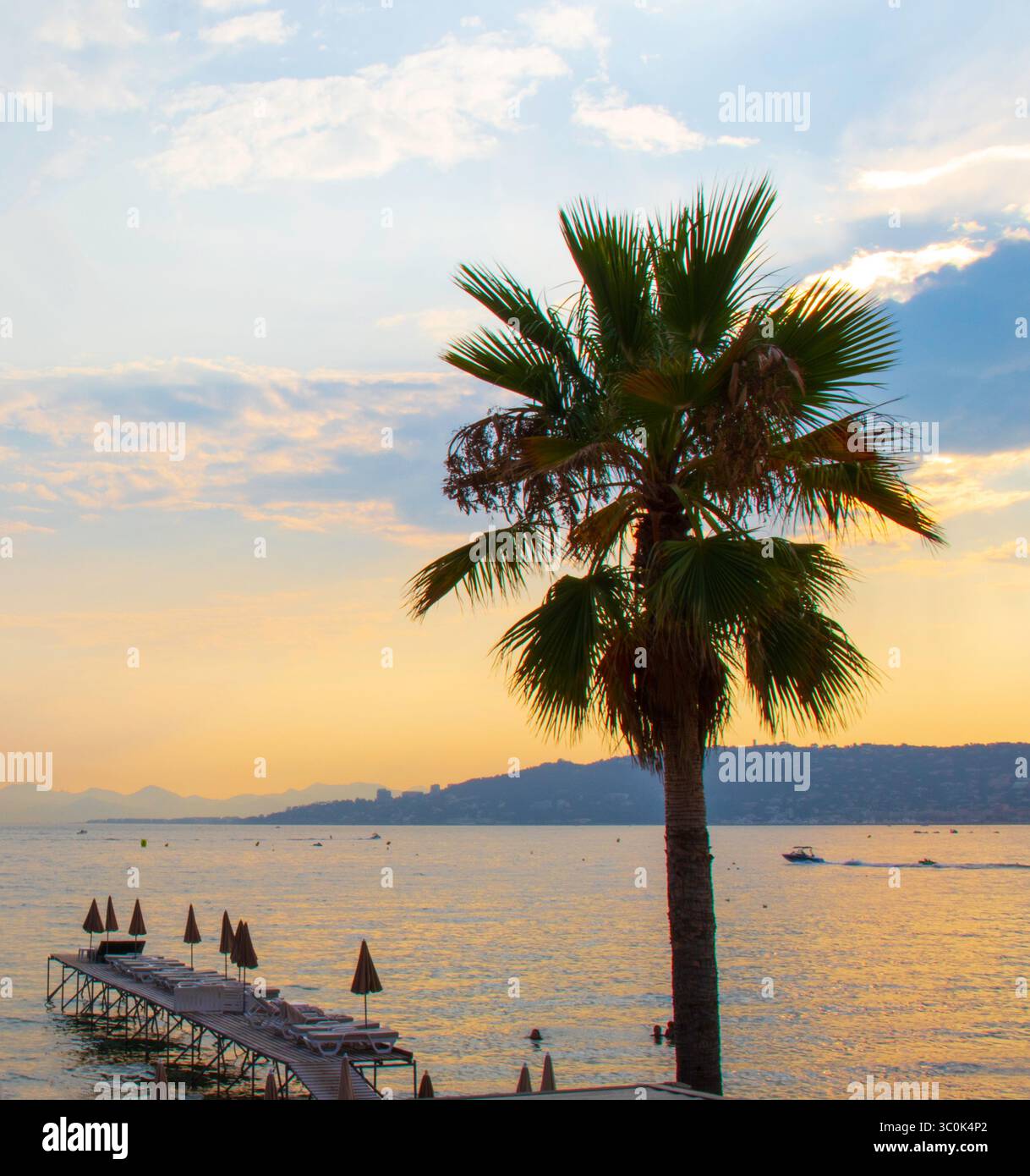 Coucher de soleil sur la côte donnant sur la mer à Juan les Pins sur la Côte d'Azur dans le sud de la France. Avec un palmier, une jetée et des collines lointaines. Banque D'Images
