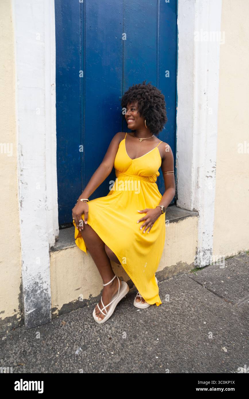 Belle jeune femme aux cheveux afro, vêtue d'une robe jaune, assise près d'une porte bleue d'une vieille maison. Personne confiante. Brésil Banque D'Images