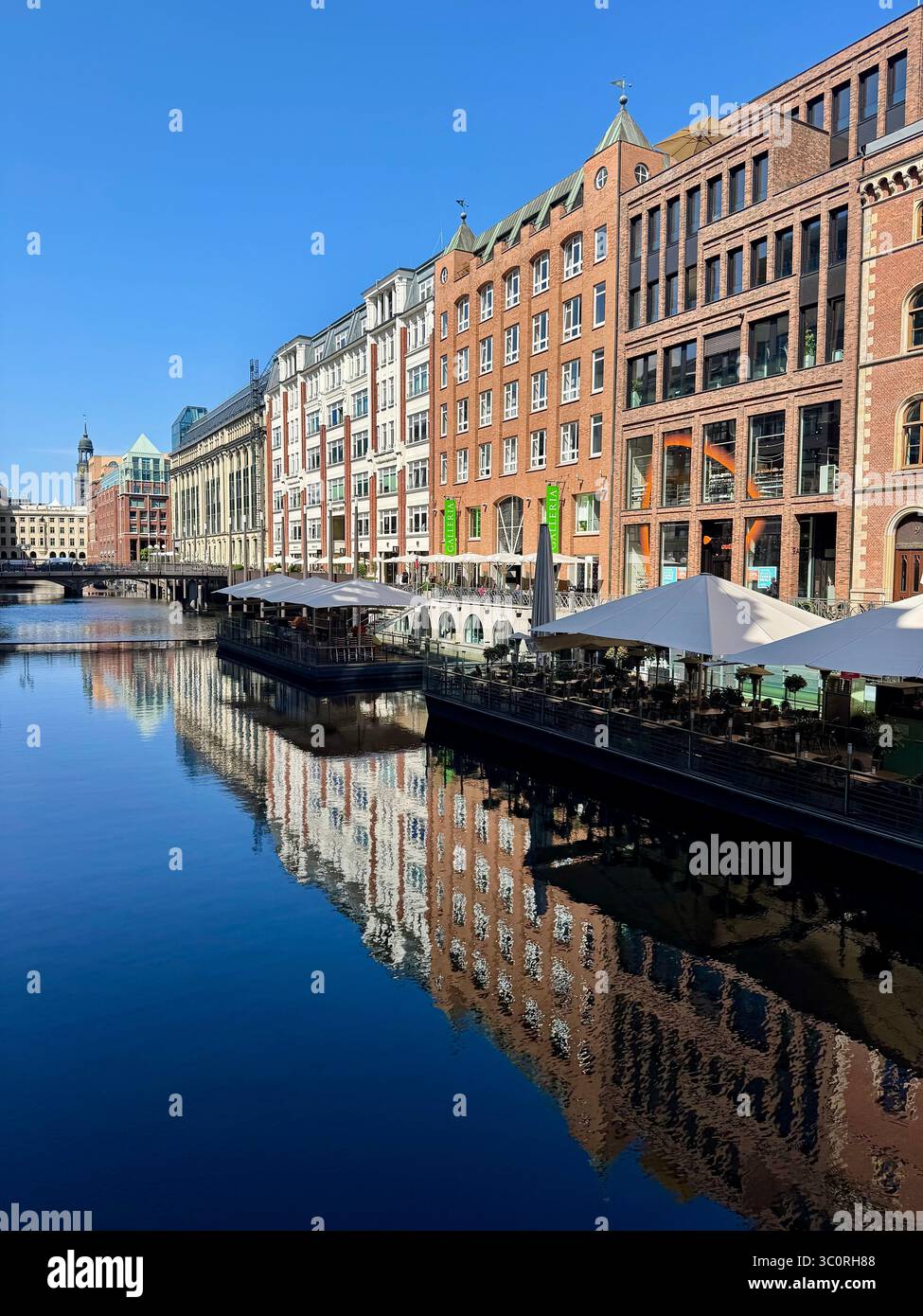 Bâtiments historiques et modernes le long du canal avec des cafés en plein air et leurs reflets dans l'eau lors d'une journée de ciel bleu clair à Hambourg, en Allemagne. - Image de stock capturée avec un smartphone