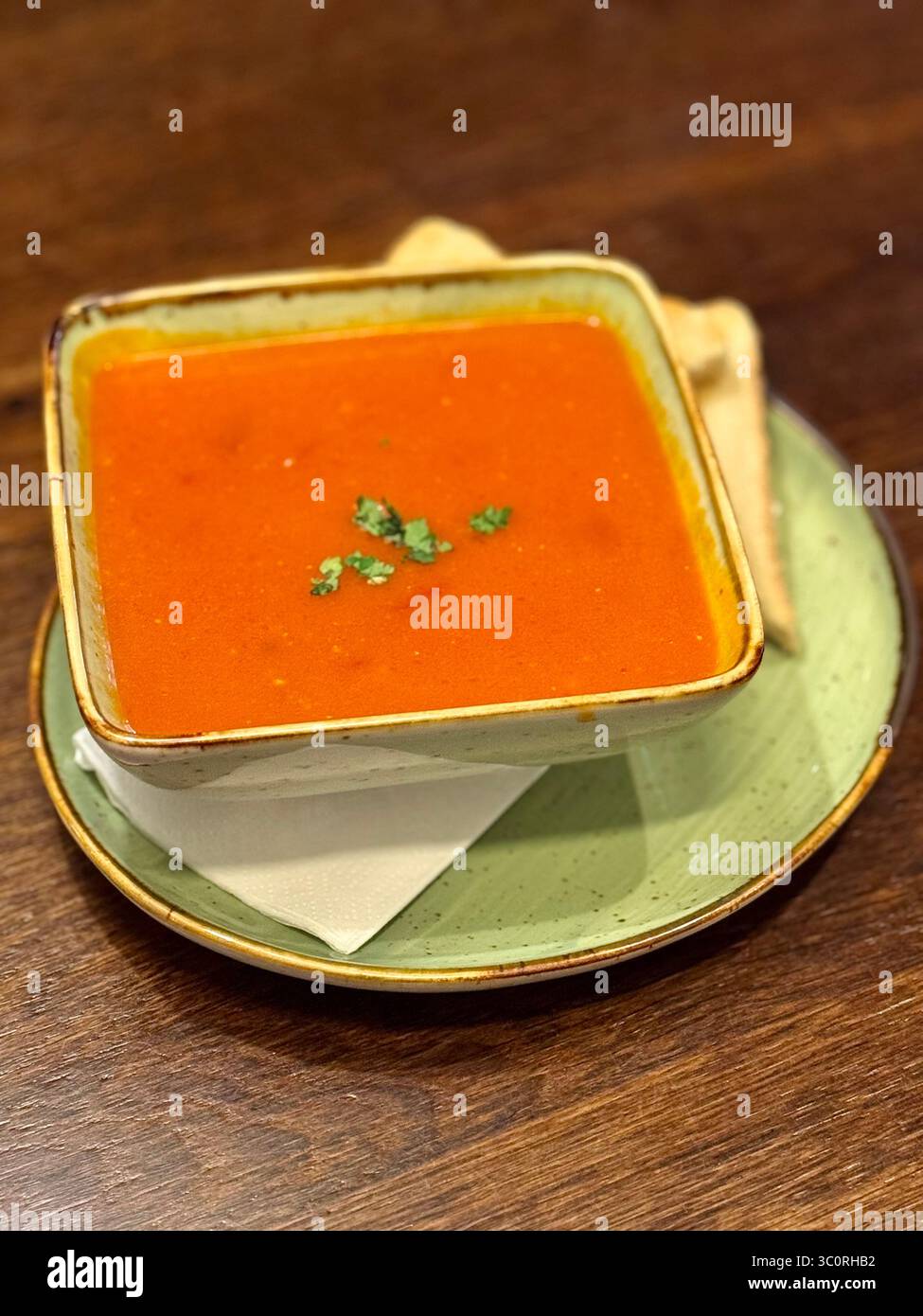 Bol de soupe chaude aux tomates garnie de persil, servi avec du pain grillé sur une table en bois rustique. - Image de stock capturée avec un smartphone